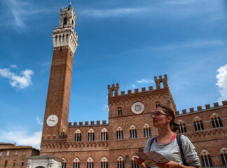 Fototapeta premium Siena, Tuscany, Italy. August 2020. A woman in piazza del campo, on the right the tower of the Mangia: symbol of the city. She consults the map and looks around