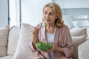 Beautiful elderly woman eats her vegetable salad sitting at home on the couch