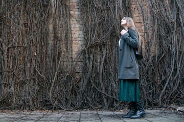 Young woman in a coat stands against the backdrop of a wall of withered ivy. Side view.