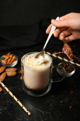 Woman stirring tasty almond latte in glass cup on dark background