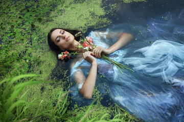 Art beautiful romantic woman lies in swamp in blue long dress with flowers. Portrait brunette in transparent dress in water swamp mud duckweed. Book cover