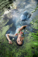 Fototapeta premium Art beautiful romantic woman lies in swamp in blue long dress with flowers. Portrait brunette in transparent dress in water swamp mud duckweed. Book cover
