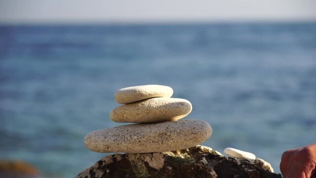Man bilds stones pyramid on the seashore on a sunny day on the blue sea background. Happy holidays. Pebble beach, calm sea, travel destination. Concept of happy vacation on the sea, meditation, spa
