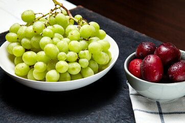 Branch of ripe green grape on plate with water drops and plums in bowl. Juicy fruits on wooden background, closeup. Healthy food on dark kitchen table