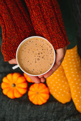A woman in a brown knitted sweater and yellow socks is holding a cup of cappuccino coffee against a gray blanket. A warm and cozy autumn. Hugge. Peace and comfort. Yellow-orange pumpkins. Autumn mood