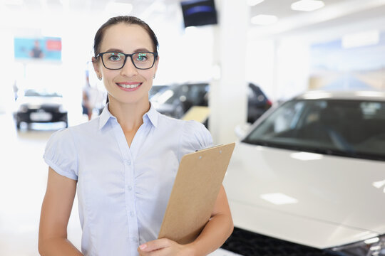 Young smiling woman manager in glasses holds clipboard on background of car dealership closeup