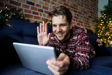 Smiling man waving hand to video call through digital tablet in living room