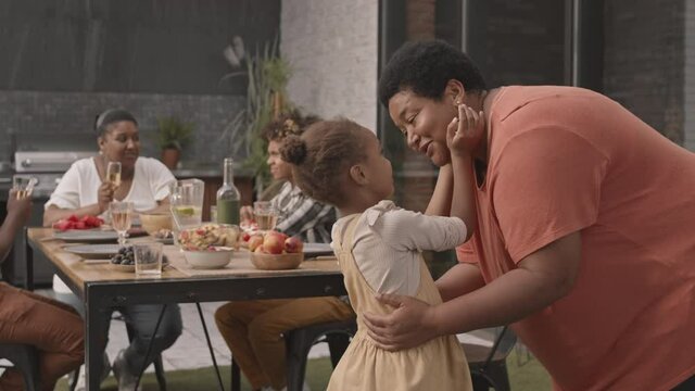 Medium long of happy African grandmother talking to and hugging her adorable little granddaughter in foreground of other family members dining at table on patio