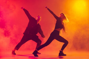 Bottom view of two young hip-hop dancers in motion isolated over gradient pink yellow background in neon light in smoke