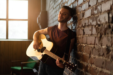 Man with acoustic guitar leaning against brick wall playing music singing songs enjoy life Medium...