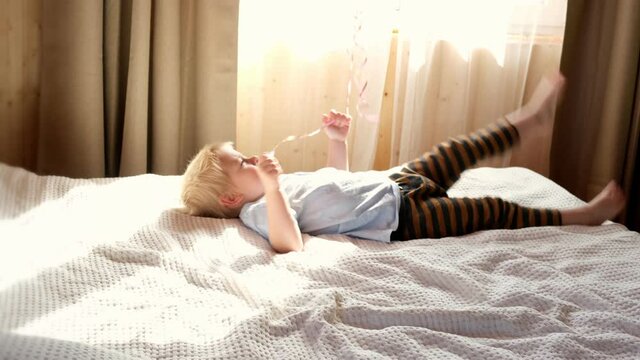 Cute little boy in striped clothes jumping on bed in cozy sunny bedroom. Child play with balloons at home enjoy carefree time. Kid have fun and laughing on quarantine, coronavirus home isolation.