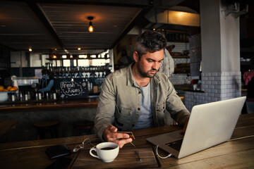 Stressed caucasian business man making online payment with card while working on laptop drinking coffee in trendy cafe.