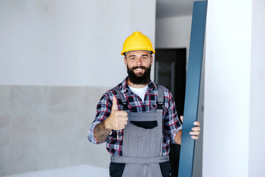 A Happy, Smiling, Construction Site Worker Is Standing In A New Building With A Metal Beam In Hand. He Is Showing Thumbs Up And Looking At The Camera. 