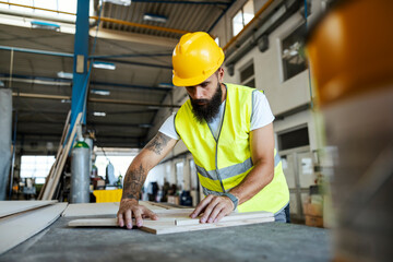 A bearded tattooed worker in a protective uniform is working with wooden parts in his workshop. Woodwork in workshop