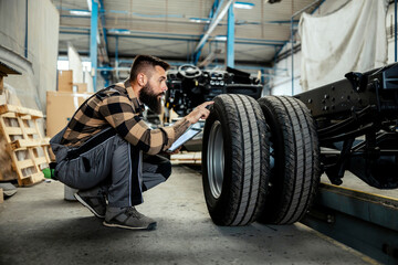 Fototapeta premium An auto-mechanic crouching next to a tire with a tablet in his hands and checking on it. Worker in the auto-mechanic workshop