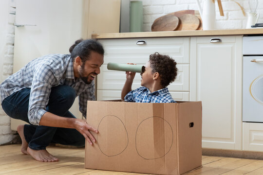 Happy African American father with little son playing with cardboard box and toy spyglass in kitchen, loving young dad and 5s boy child engaged in funny activity, spending leisure time at home