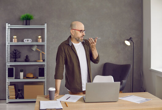 Modern technology. Businessman answers incoming call or leaves voice audio message to customer remotely. Busy serious middle-aged man in his office communicates by phone using loudspeaker mode.