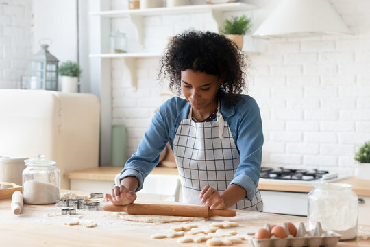 African American woman rolling out dough, cooking homemade cookies or pie at home, attractive young female housewife baker wearing apron baking pastry, using rolling pin, standing in cozy kitchen