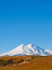 Fototapeta premium A beautiful asphalt road through the autumn hills of the Caucasus leads to Elbrus. Elbrus with two peaks against the blue sky is covered with snow. Copy space.