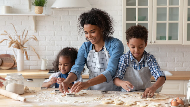 Loving African American mother with kids cooking homemade cookies together, using dough cutters, caring young mom with little son and daughter wearing aprons spending leisure time in kitchen at home - Powered by Adobe