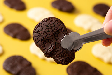 Hand picking up chocolate Mexican skull with tongs on yellow background