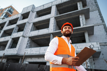 Young man engineer in workwear standing in construction site with clipboard