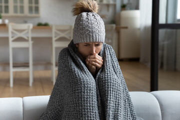 Unwell millennial female renter in hat and blanket sit in cold living room suffer from air...