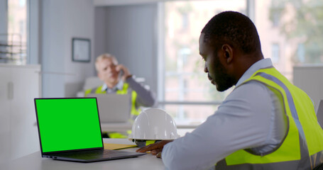 Side view of afro engineer in reflective vest looking at laptop screen in office. Green screen