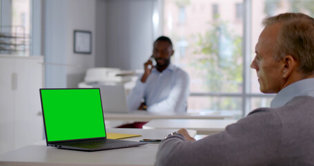 Mature businessman sitting at desk and looking at blank green screen on laptop