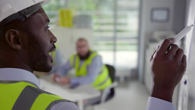 Back view of afro-american engineer in hardhat and vest writing on board at corporate meeting