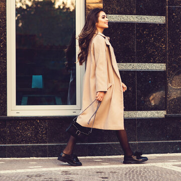 Young stylish woman walking in street. Woman wearing trendy coat, fashion shoes and holding black handbag. Fashion female blooger going shopping