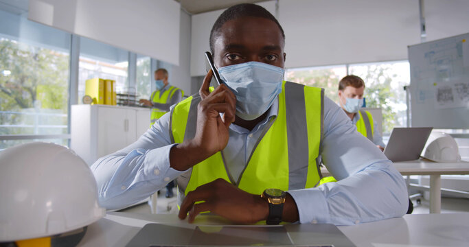 Afro-american engineer in safety mask sitting at desk in office and talking on smartphone