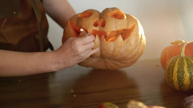 Pumpkin carving by female hands in apron as autumn seasonal Halloween holiday tradition. Woman carves a mouth in a pumpkin to make a jack o'lantern on a wooden table. High quality image