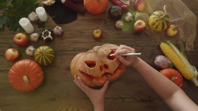 Halloween concept: top view of female hands carving pumpkin. Woman carves an eye in a pumpkin to make a jack o'lantern on a wooden table with vegetables and candles. High quality image