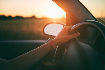 Female hands of car driver on steering wheel, road trip at sunset