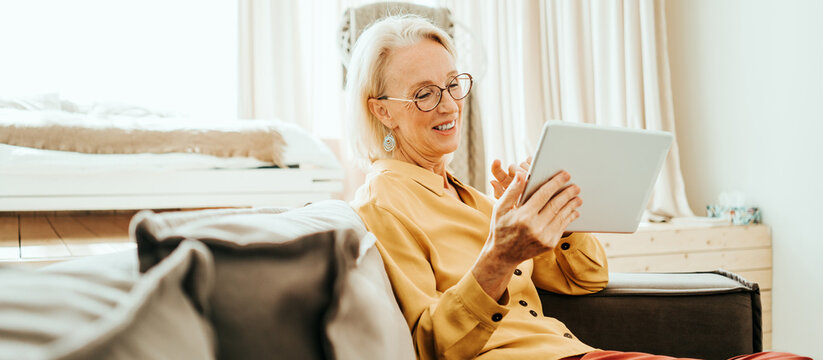 Handsome mid aged woman at her bright and sunny apartment. Happy and smiling using tablet computer