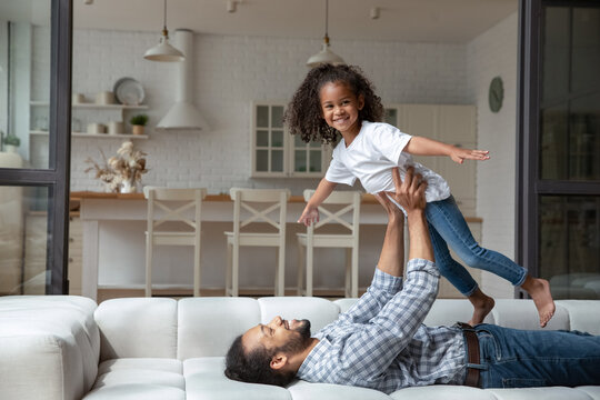 African American father lying on couch, lifting happy little daughter looking at camera, pretending flying with hands outstretched, loving dad and 5s girl child engaged in funny activity at home