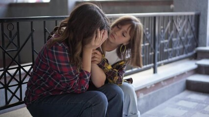 Beautiful nerd Caucasian teenage girl in eyeglasses sitting outdoors crying as empathetic classmate coming hugging calming down friend. Depressed stressed bullied student supported by friend