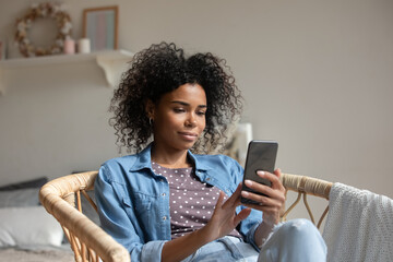 African American woman using smartphone, relaxing sitting in cozy armchair, attractive young female...