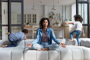 Concentrated African American woman meditating, practicing yoga, ignoring noisy little kids jumping...