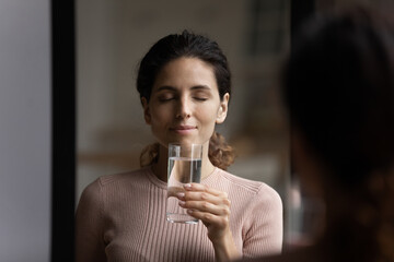 Happy millennial Caucasian female hold glass of pure mineral still water feeling thirsty dehydrated. Smiling calm young woman enjoy drink clean clear aqua follow healthy lifestyle. Hydration concept.