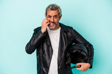 Middle age biker caucasian man holding helmet isolated on blue background  biting fingernails,...