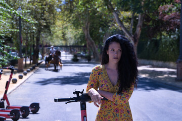 Middle-aged adult Hispanic woman with black curly hair, wearing a colorful outfit, leaning on an electric scooter. Concept scooter, urban mobility, electricity, battery, ecology.