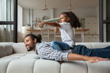 Happy African American girl sitting on father back, playing funny game, holding cardboard tube as...