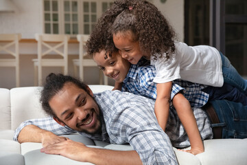 Funny African American little kids lying on smiling loving father back, caring young dad with cute little 5s son and daughter having fun together, enjoying tender moment, relaxing on couch at home