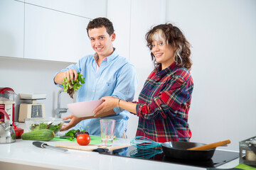 pareja joven sonriente hace una ensalada en un bol rosa y pica tomate sobre una tabla de cortar...