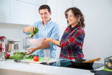 pareja joven sonriente hace una ensalada en un bol rosa y pica tomate sobre una tabla de cortar...