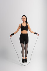 full length of cheerful sportswoman exercising with skipping rope on grey