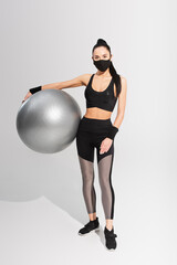 Fototapeta premium young sportswoman in sportswear and black protective mask holding fitness ball on grey