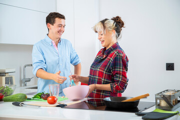 mujer joven sonriente bate huevos en un bol rosa con una varilla de cocina azul mientras rie con su pareja en una cocina blanca con verduras y vegetales